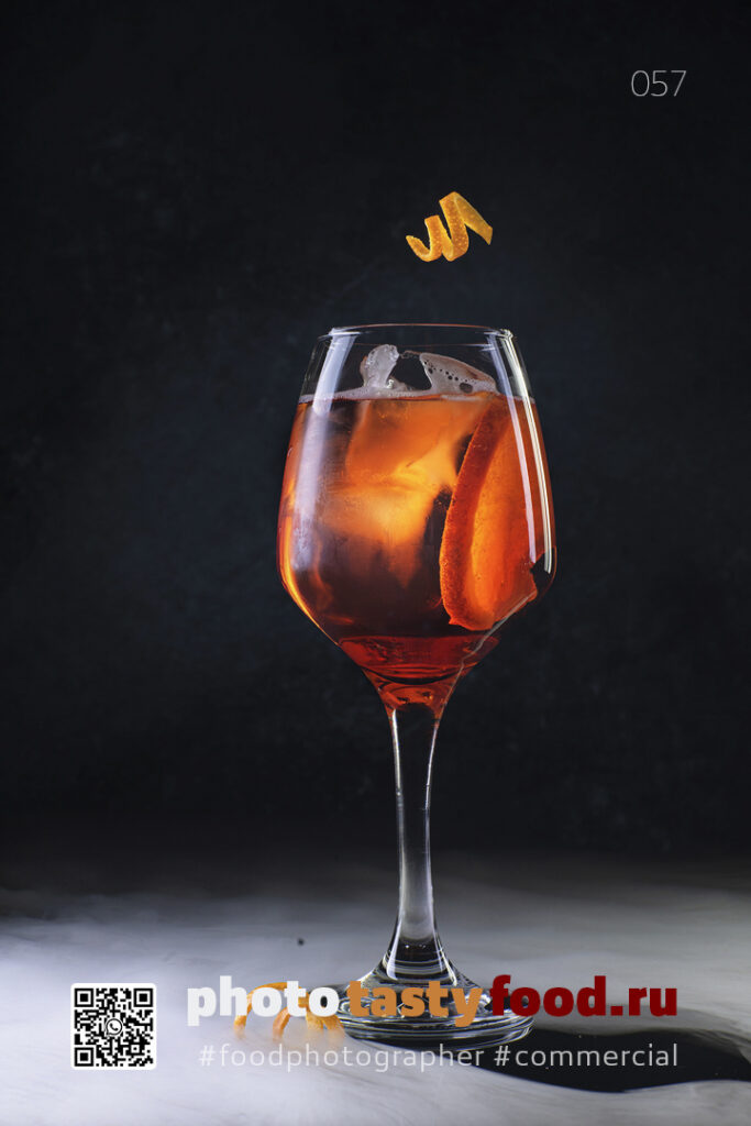 Освежающий Aperol Spritz в бокале для вина, украшенный долькой апельсина и завитком, на темном фоне. A refreshing Aperol Spritz in a wine glass, garnished with an orange slice and a twist, set against a dark background. Фуд фотограф | food photographer